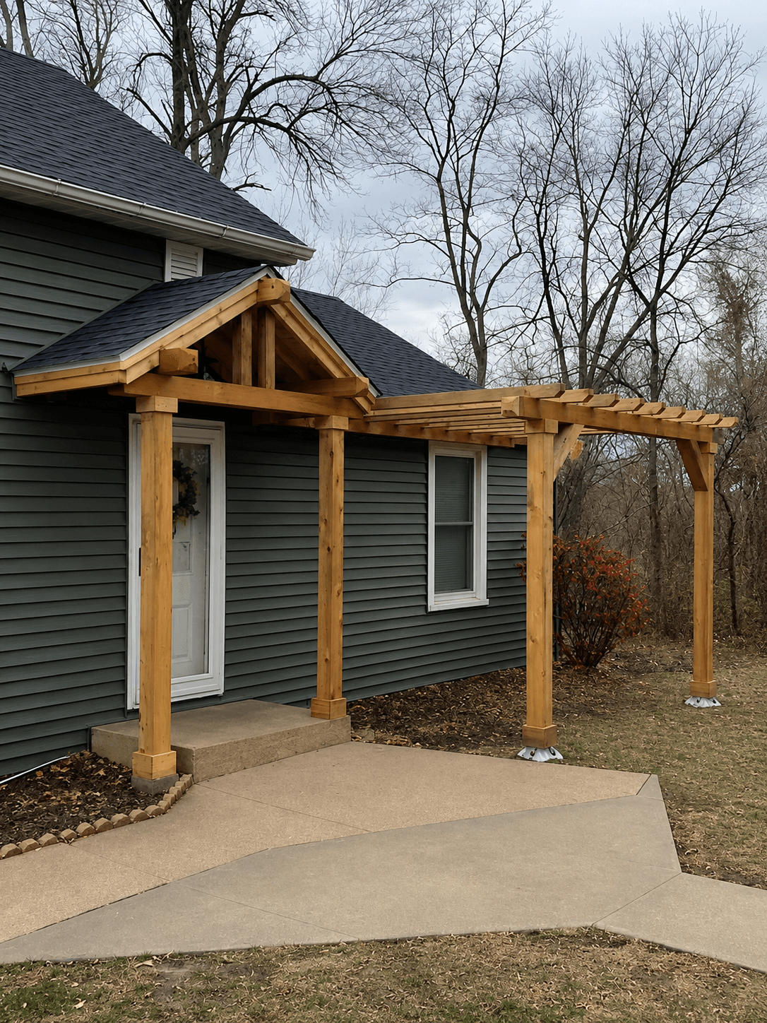 New wooden gabled porch and pergola attached to a dark grey house with concrete walkway.
