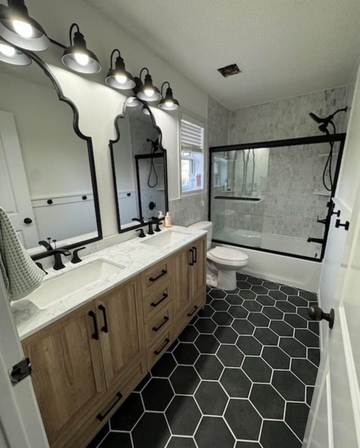 Modern bathroom featuring double wood vanity, black arched mirrors, and black hexagonal floor tiles.