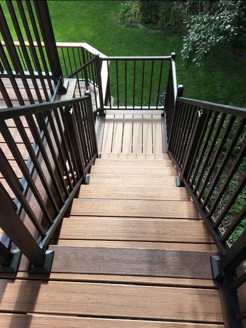 Brown composite deck stairs with black metal railings descending toward a lush green lawn.