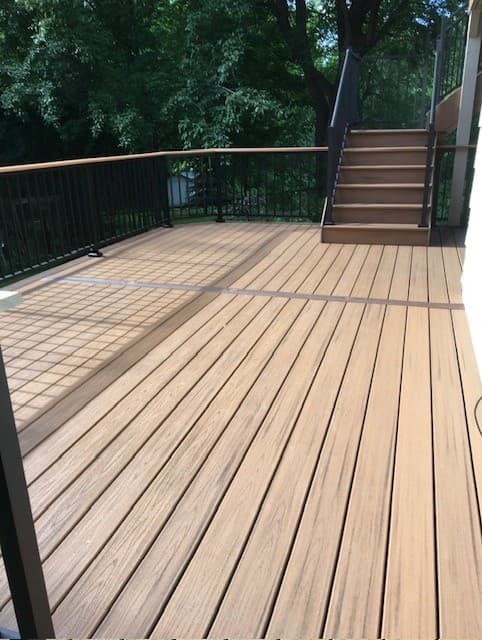 Modern tan deck with black metal railings and stairs, surrounded by lush green trees.