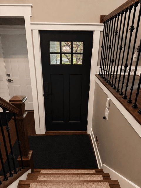 Black front door with six-pane window and white trim, viewed from a wooden staircase.