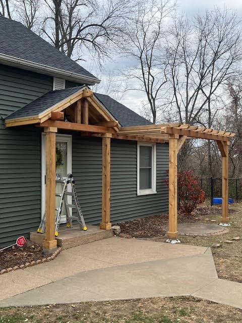 Wooden gabled porch and pergola attached to a dark grey house with a ladder.