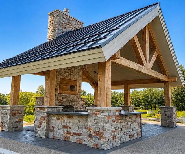 Timber frame outdoor kitchen pavilion with stone masonry, a chimney, and black metal roof.