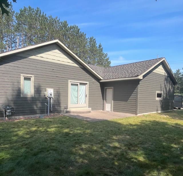 Gray-sided house with sliding glass door and patio, set against green lawn and pine trees.
