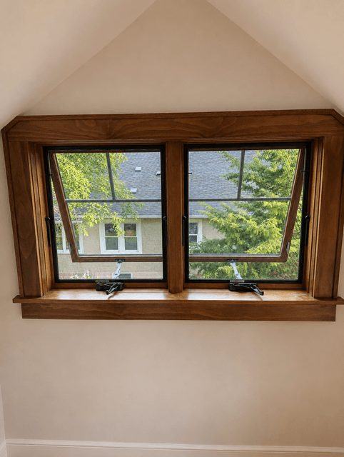 Two wooden windows tilted open, showing a view of green trees and a neighboring house.